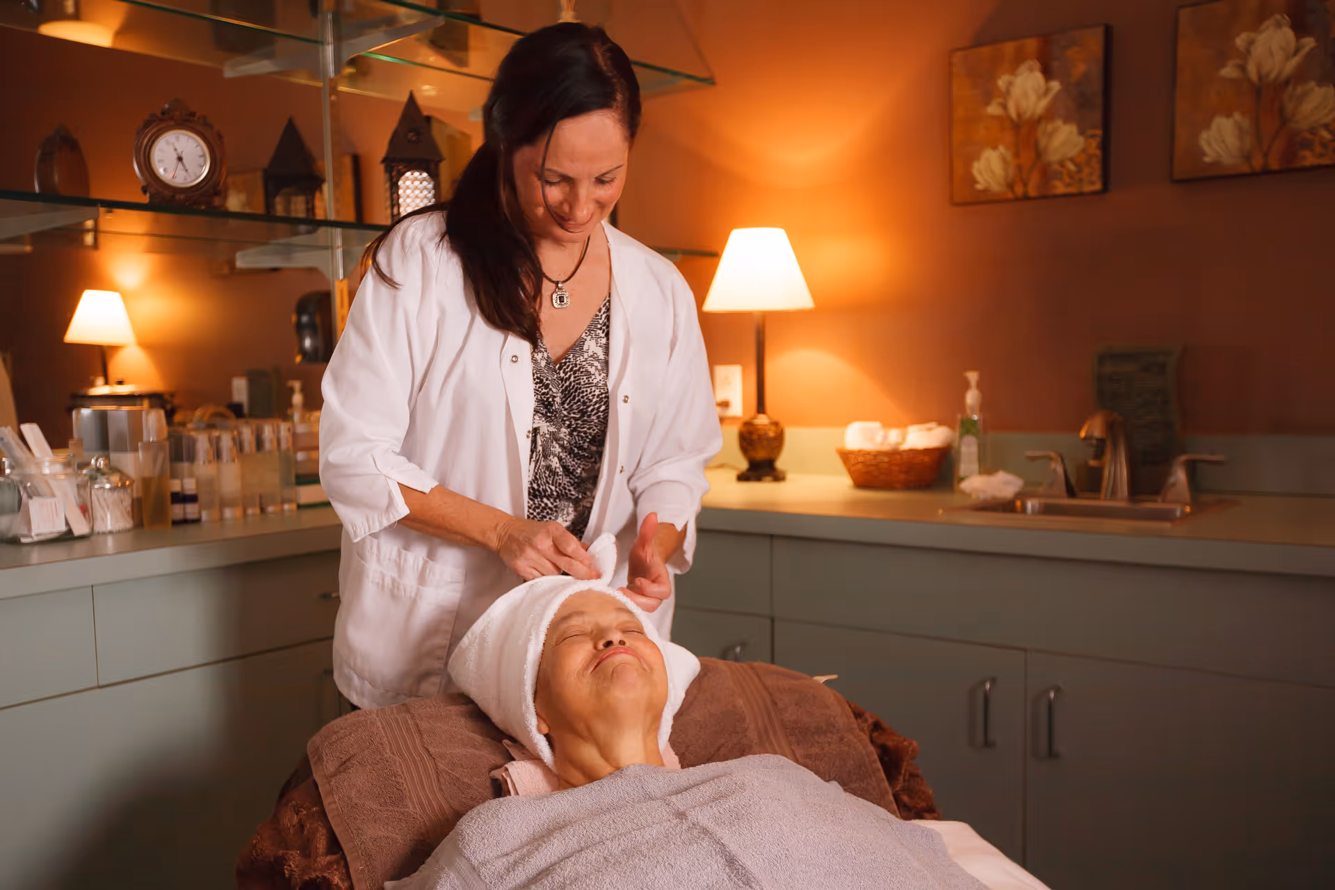A woman in a white coat is gently wrapping a towel around the head of an elderly woman lying on a treatment bed in a warmly lit room with soft brown walls, a lamp, and floral paintings on the wall.
