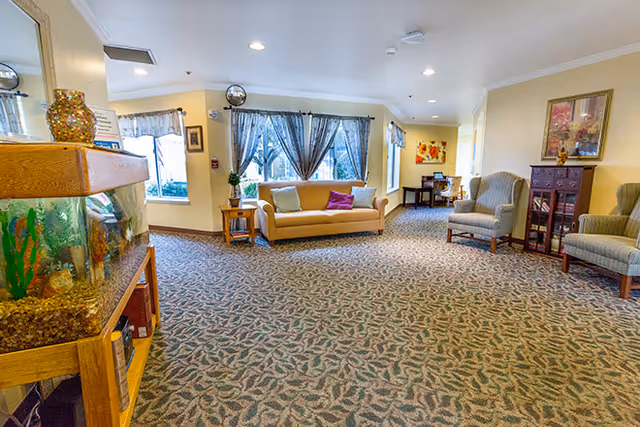 A spacious living room area in a senior living facility with patterned carpet, a beige sofa with colorful pillows, two armchairs, a wooden side table with a plant, a fish tank on a wooden stand, and large windows with curtains allowing natural light to fill the room. There are framed pictures on the walls and a small table with chairs in the background.