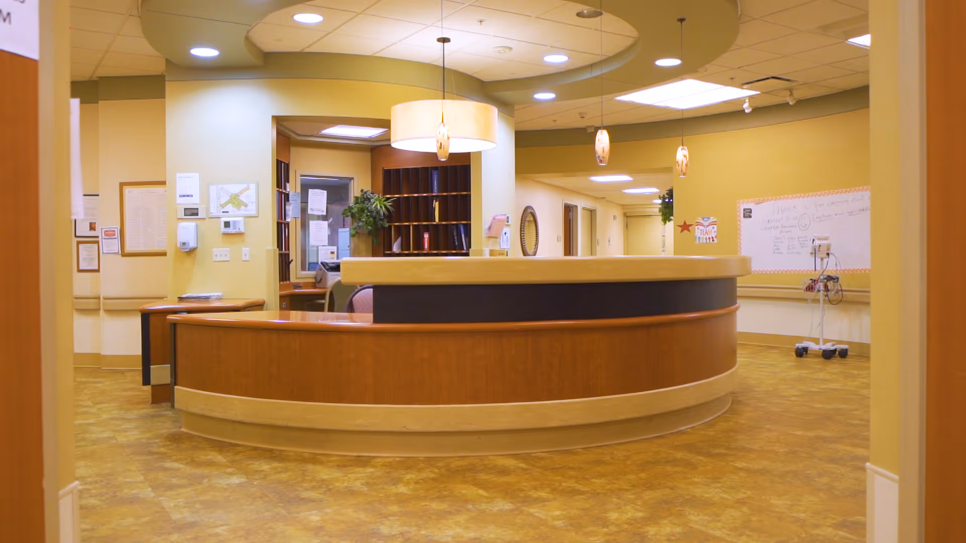 Reception area inside Medilodge of Capital Area featuring a curved wooden front desk with a beige countertop, overhead pendant lights, yellow walls, and a hallway leading to other rooms. There is a whiteboard on the right wall and some office equipment behind the desk.