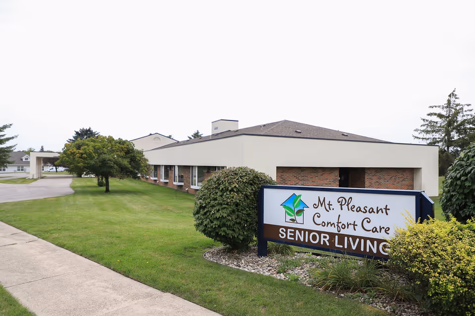 Exterior view of Mt. Pleasant Comfort Care Assisted Living and Memory Care building with a sign in the foreground that reads 'Mt. Pleasant Comfort Care Senior Living'. The building is single-story with a beige and brick facade, surrounded by green grass, bushes, and trees under a cloudy sky.