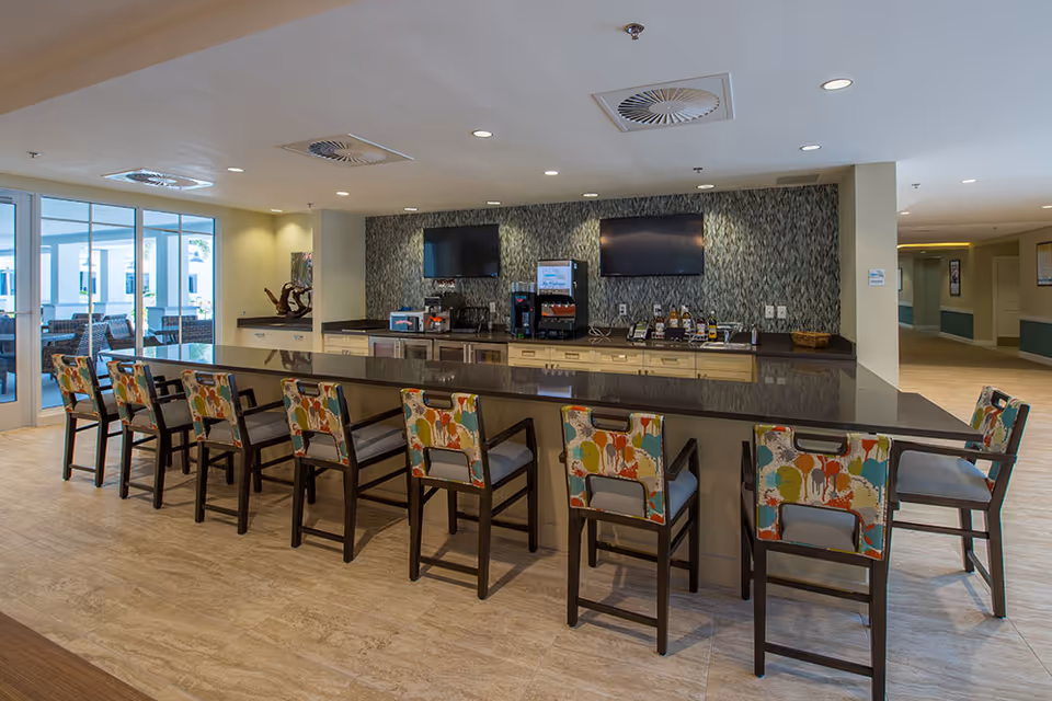 A modern communal kitchen area with a long counter and eight high chairs featuring colorful patterned upholstery. The back wall has two flat-screen TVs mounted above a countertop with various beverage dispensers and coffee machines. The room has large windows on the left side letting in natural light and a hallway visible on the right.
