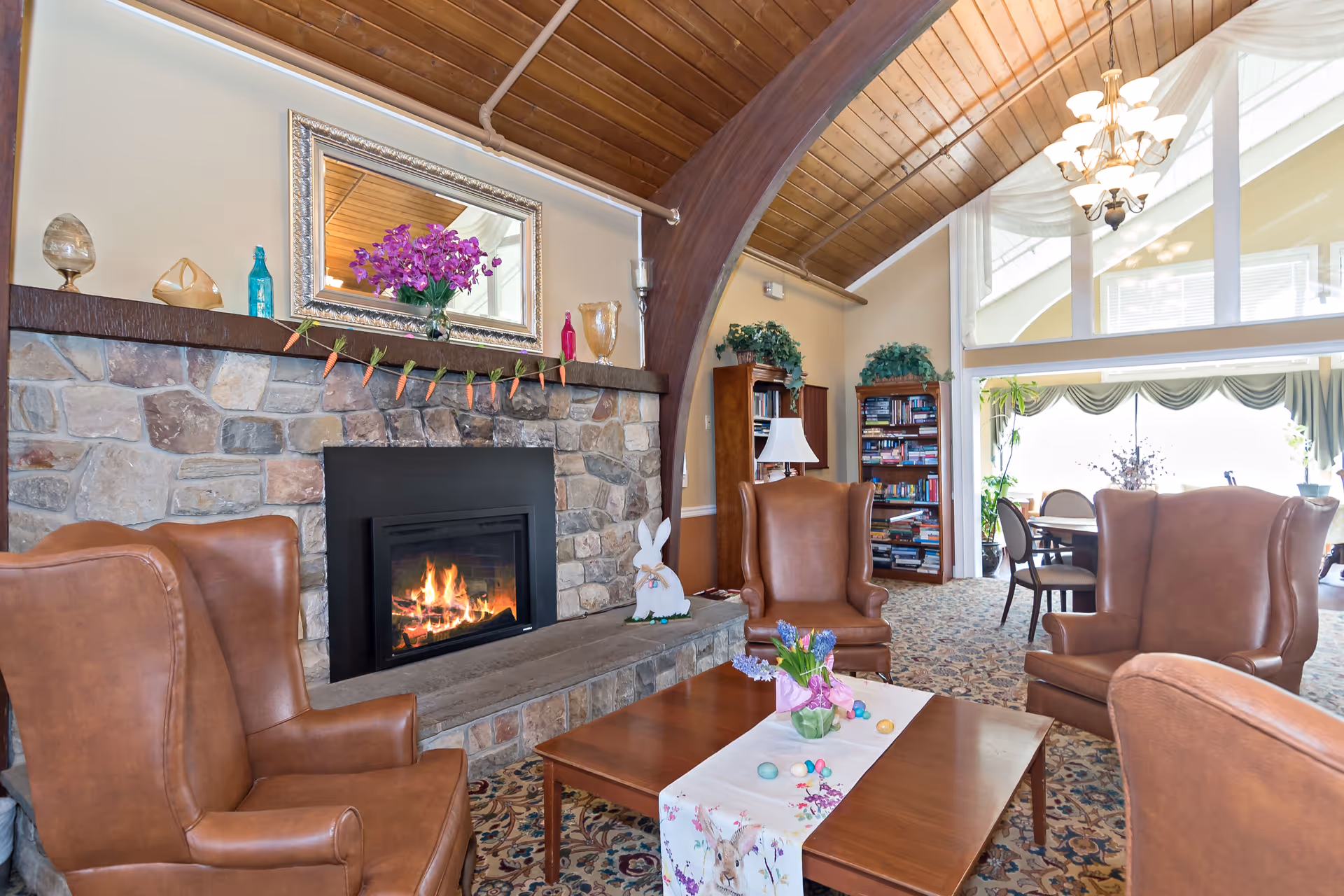 A cozy living room area with a stone fireplace featuring a lit fire, decorated with a mirror, flowers, and small ornaments. Four brown leather armchairs surround a wooden coffee table with a floral table runner and Easter-themed decorations. The room has a high wooden ceiling with a chandelier, bookshelves filled with books, and large windows letting in natural light.