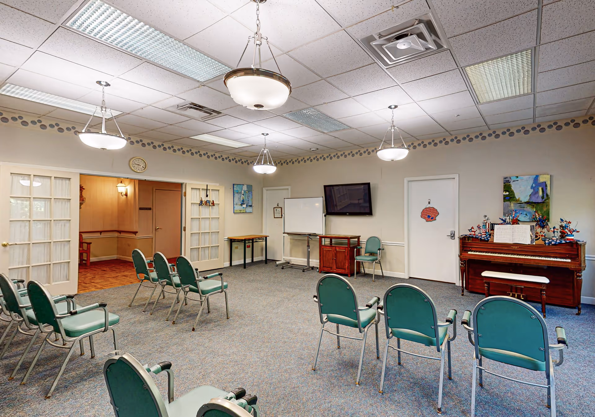 Spacious community activity room with rows of green chairs facing a piano, a wall-mounted TV, and a whiteboard.