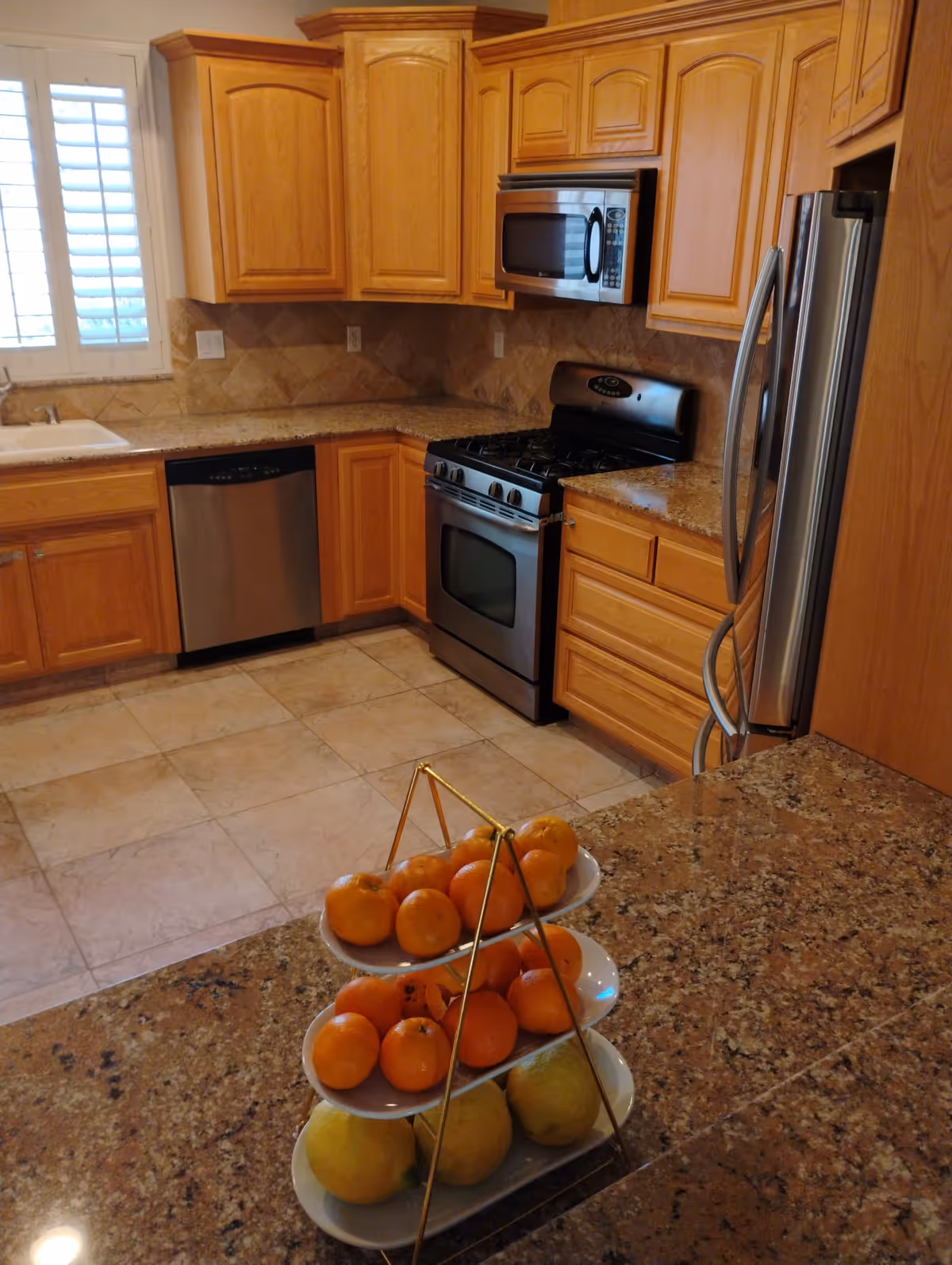 A kitchen with wooden cabinets, granite countertops, and tiled floor. The kitchen includes a stainless steel refrigerator, stove with oven, microwave, and dishwasher. A three-tiered fruit stand with oranges and other citrus fruits is placed on the countertop in the foreground. A window with white shutters is visible above the sink.