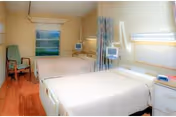 A clean and well-lit patient room in a care facility featuring two hospital beds with white linens, medical monitors mounted on the wall, a green upholstered chair, a window with a view of greenery, and light-colored walls and flooring.