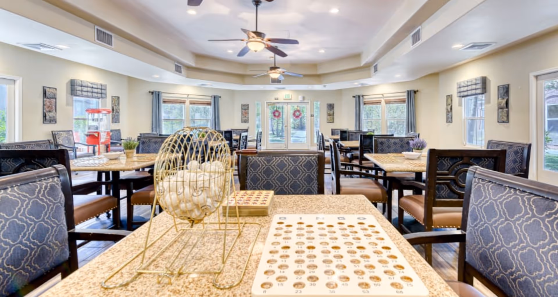 Bright communal dining/activity room with bingo equipment on a foreground table, multiple dining tables and chairs, ceiling fans, and windows.