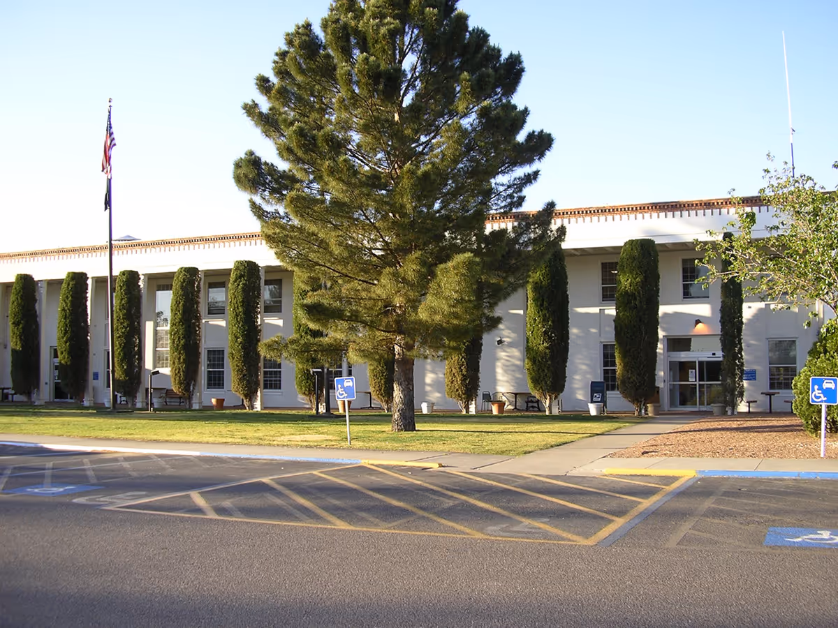 Front exterior of the New Mexico State Veterans Home building with tall trees, an American flag, and a handicapped parking area in front.