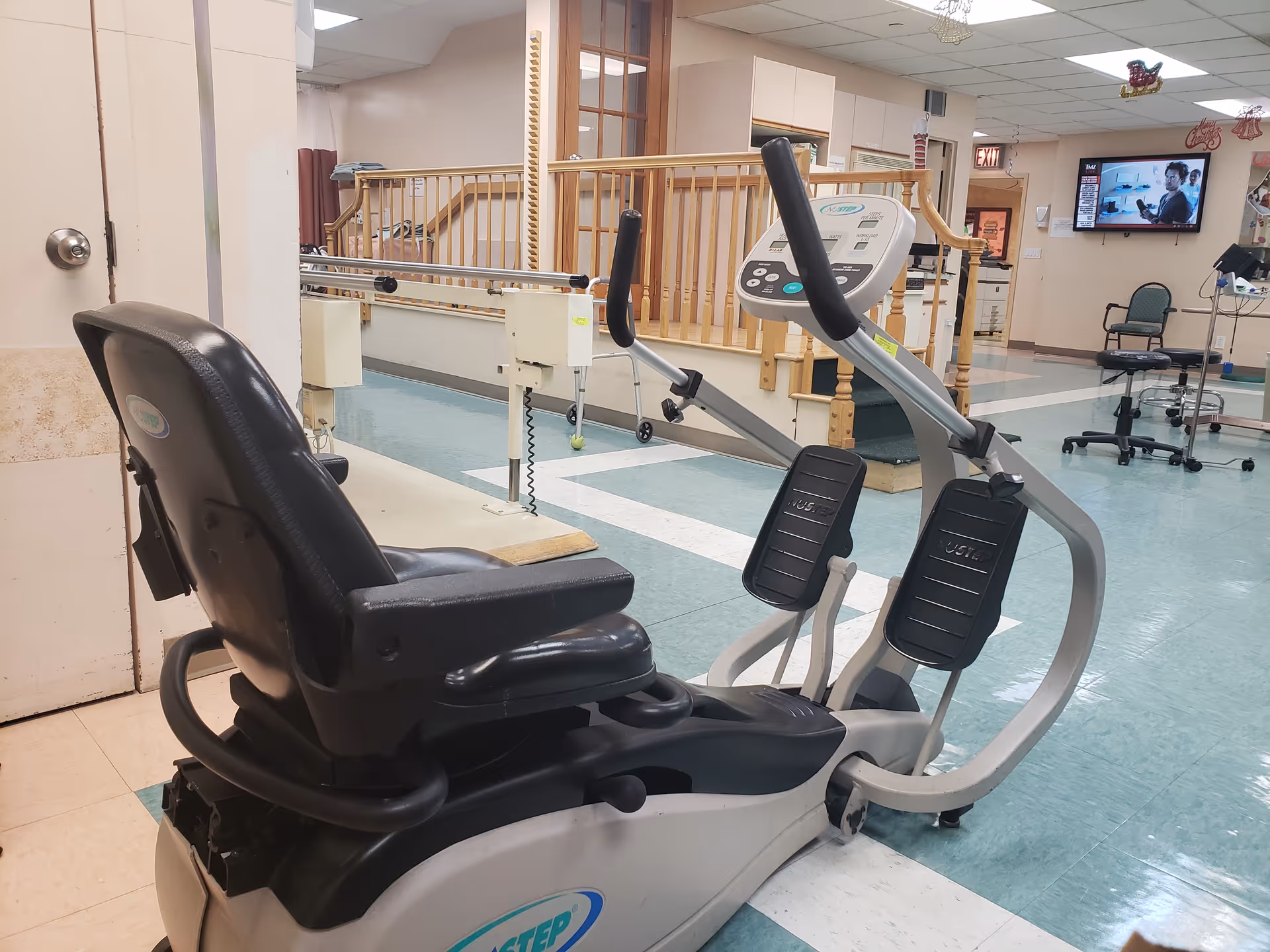 A recumbent exercise bike in a room with teal and white tiled flooring, wooden railings, and a television mounted on the wall in the background. The room appears to be part of a healthcare or rehabilitation facility with various chairs and medical equipment visible.