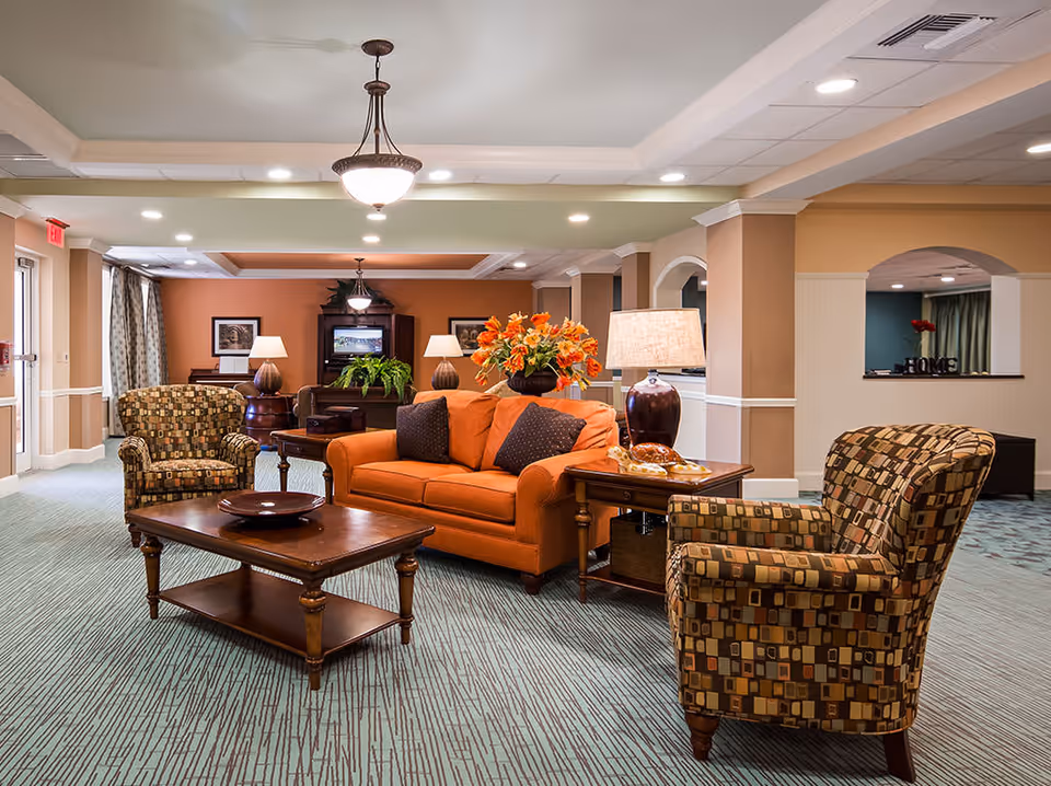 A cozy living room area in a senior living facility featuring an orange sofa with two dark pillows, two patterned armchairs, a wooden coffee table with a decorative plate, and a side table with a lamp and a flower arrangement. The room has warm beige and orange walls, carpeted floor, ceiling lights, and a television in the background.