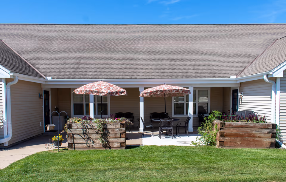 Outdoor patio area at a senior living facility with two floral-patterned umbrellas shading seating areas. There are wooden planter boxes with flowers and greenery in front of the patio. The building has beige siding and a brown shingled roof under a clear blue sky.