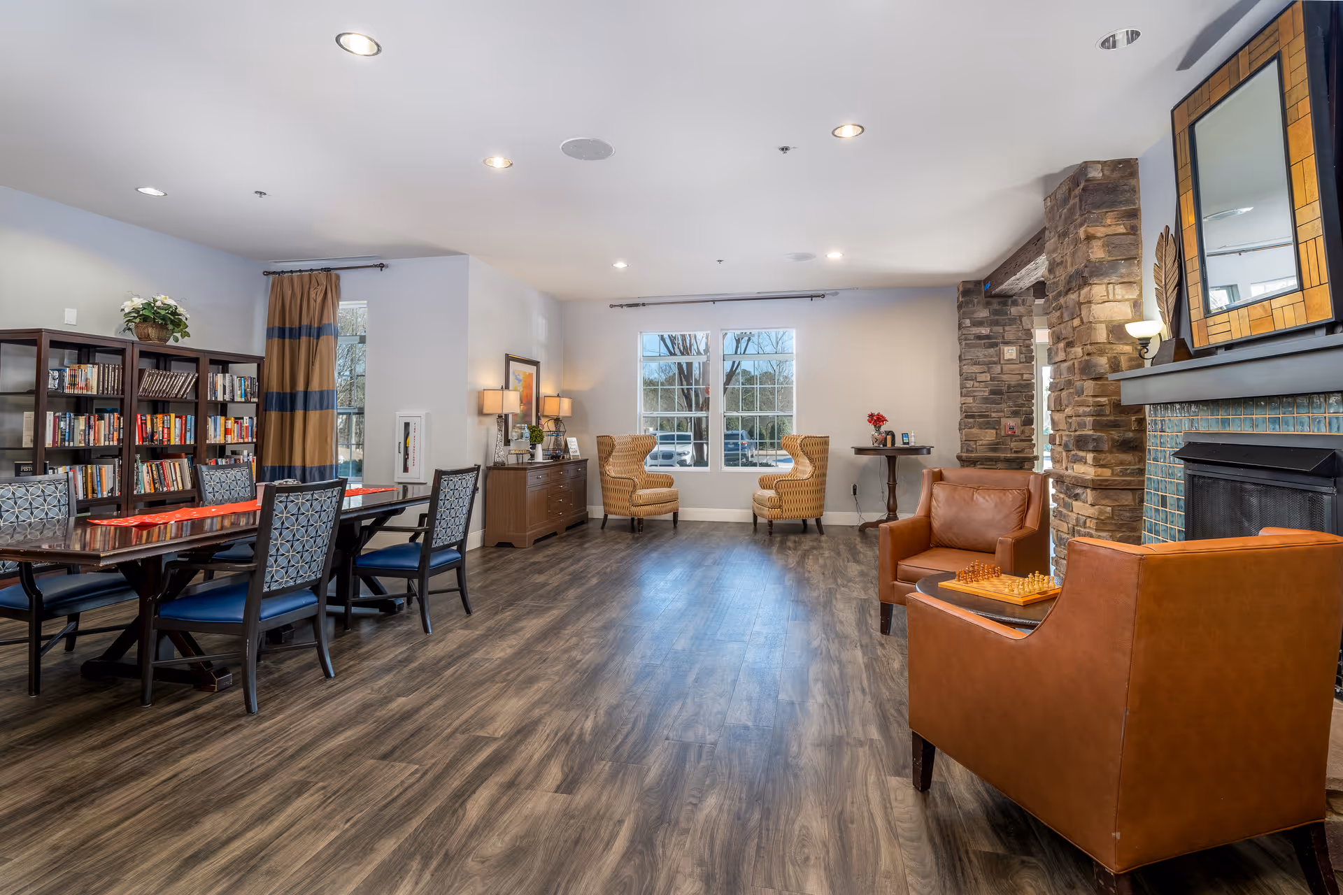 A spacious and well-lit living room area with wood flooring, featuring a fireplace with a tiled surround and a large mirror above it. Two brown leather armchairs are positioned near the fireplace with a chessboard on a small table between them. In the background, there are two patterned armchairs near a window, a wooden sideboard with lamps and framed pictures, and a bookshelf filled with books. A long dining table with chairs is also visible on the left side of the room.