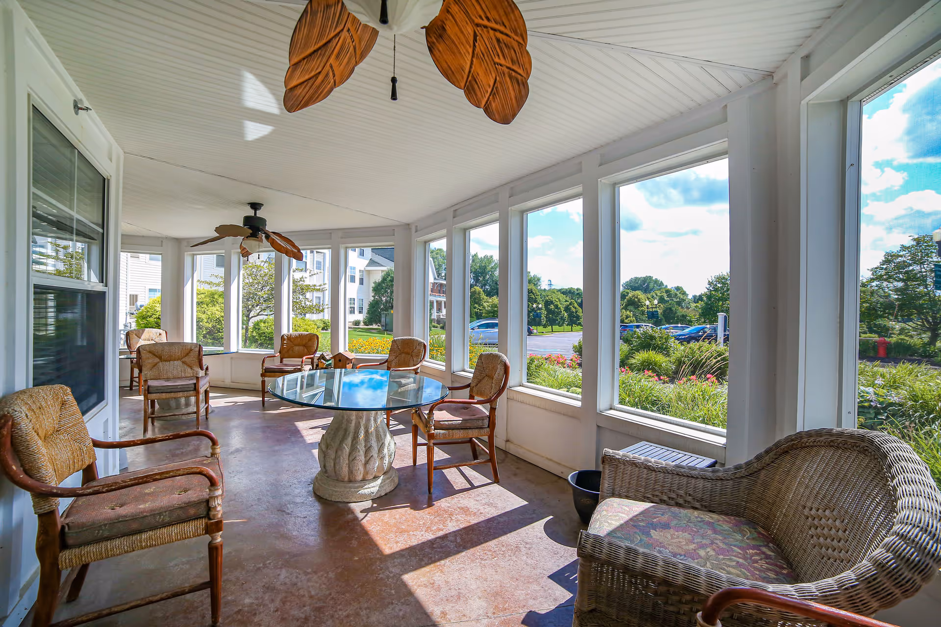 Bright sunroom with wicker chairs, a glass-top round table, ceiling fans, and large windows overlooking landscaped grounds.