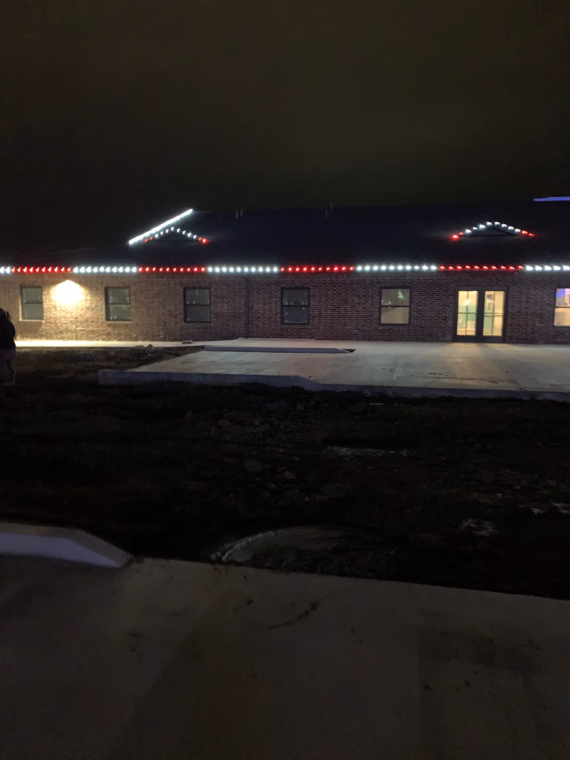 Night view of a single-story brick building with red and white lights outlining the roof. The building has several windows and a glass door entrance illuminated from inside. The surrounding area appears to be under construction or landscaping with dirt and concrete visible.