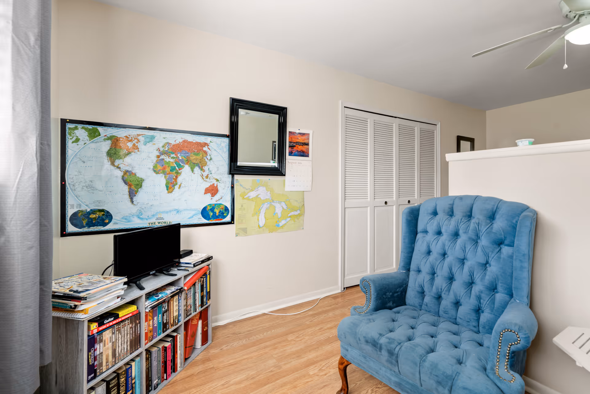 A cozy interior room with a blue tufted armchair, a bookshelf filled with books and magazines, a small TV on top of the bookshelf, and maps and a calendar hanging on the wall. The room has light-colored walls, wooden flooring, and a white closet with louvered doors.