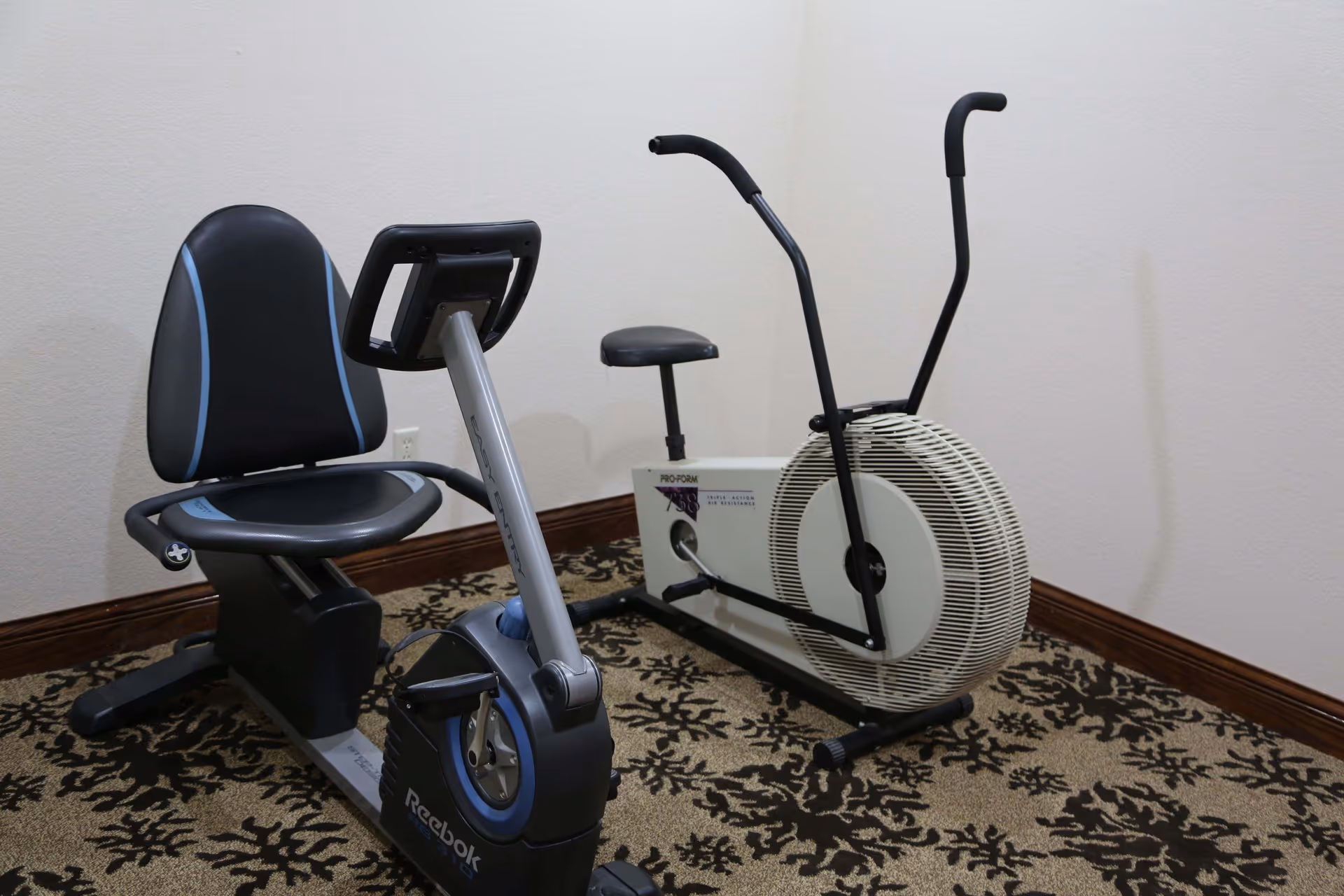 Two stationary exercise bikes positioned in the carpeted corner of an interior room with beige walls.