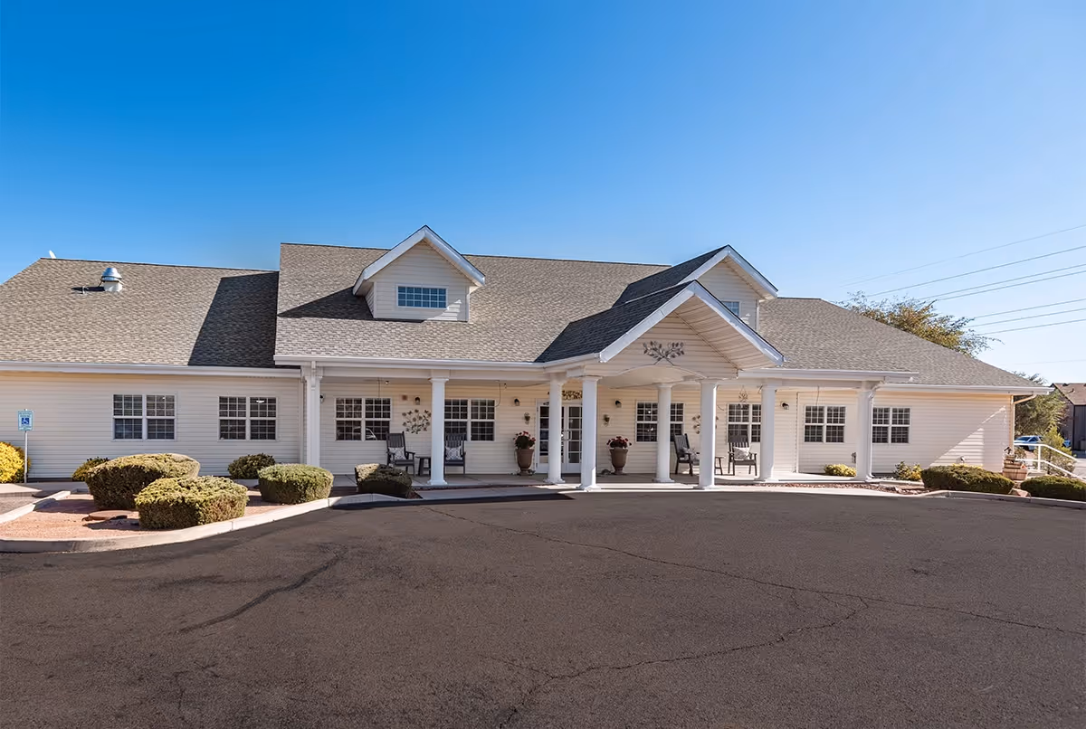 Front exterior view of a single-story senior living facility building with white siding, a covered entrance supported by white columns, several windows, and neatly trimmed bushes in front under a clear blue sky.