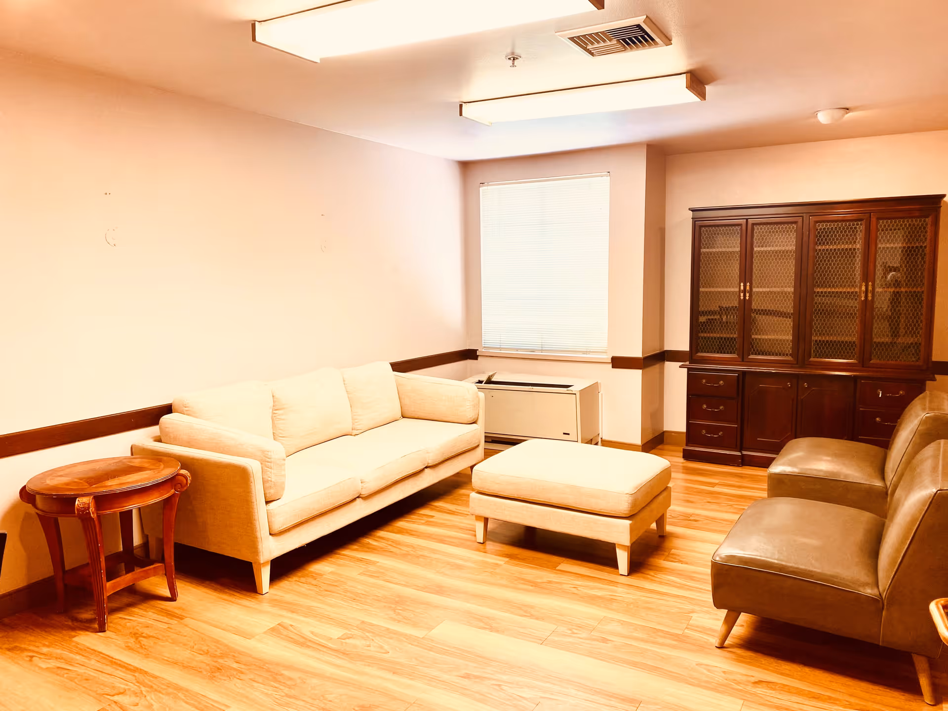 Furnished common room with a beige sofa, ottoman, leather chairs, wooden side table and cabinet on a hardwood floor.