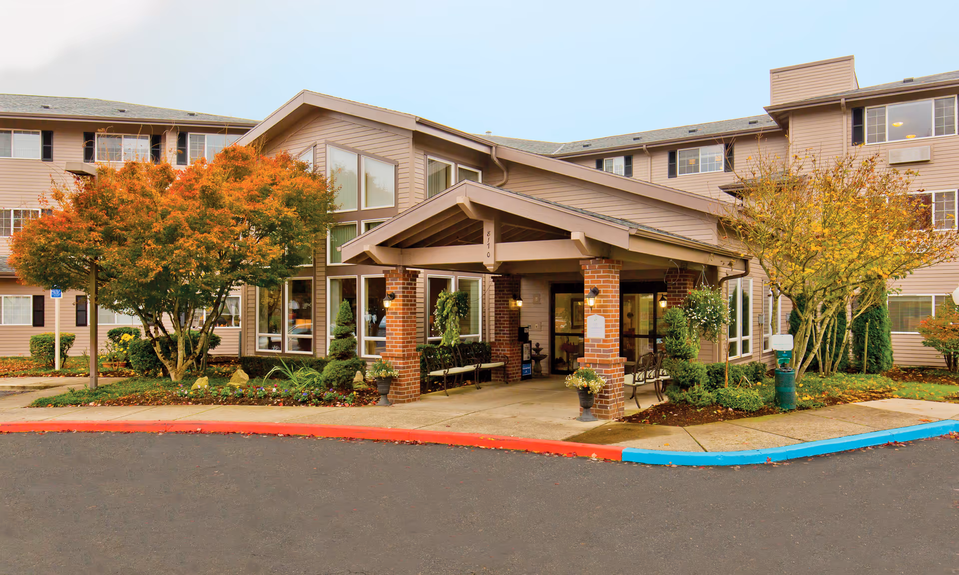 Covered entrance of a multi-story senior living building with brick columns, benches, landscaping and autumn trees.