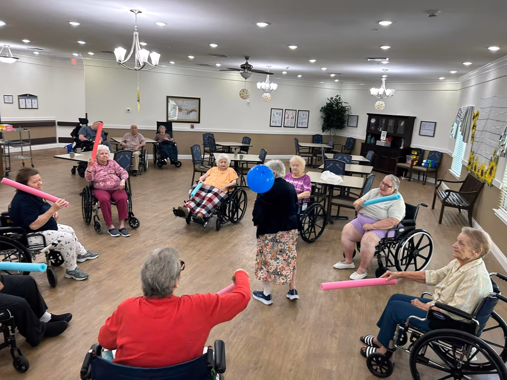 A group of elderly individuals, many in wheelchairs, participating in a seated exercise activity in a spacious, well-lit room. They are using colorful foam noodles to hit a blue balloon being held by a standing woman in the center. The room has wooden floors, several tables and chairs, chandeliers, framed pictures on the walls, and a cabinet with various items.