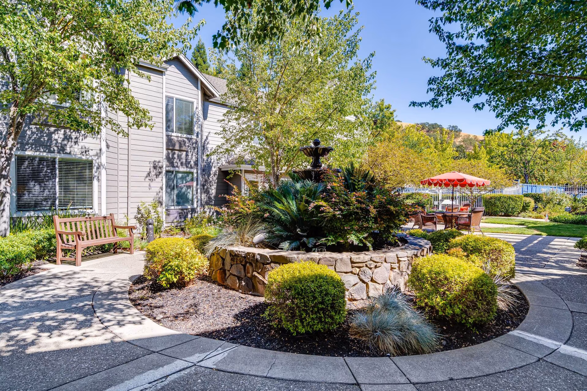 Sunny landscaped courtyard with a central stone fountain, benches, and outdoor seating with an umbrella beside a senior living building.