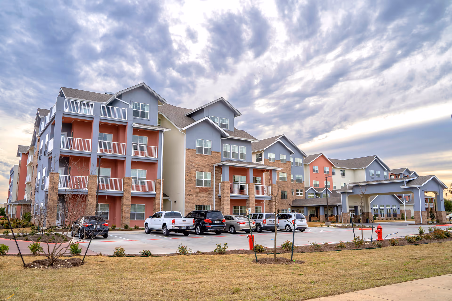 Exterior view of a multi-story senior living facility building with balconies, a parking lot with several cars, and a cloudy sky overhead. The building features a mix of brick and painted siding in muted colors, with a covered entrance area.