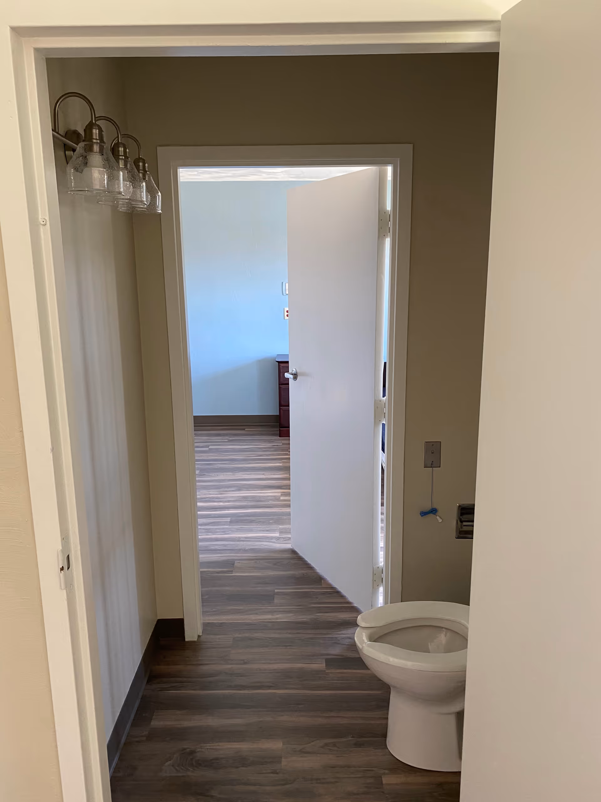 View through a bathroom doorway showing a toilet on the right, vanity light fixtures on the left, and an open door leading to a room with wood‑look flooring.