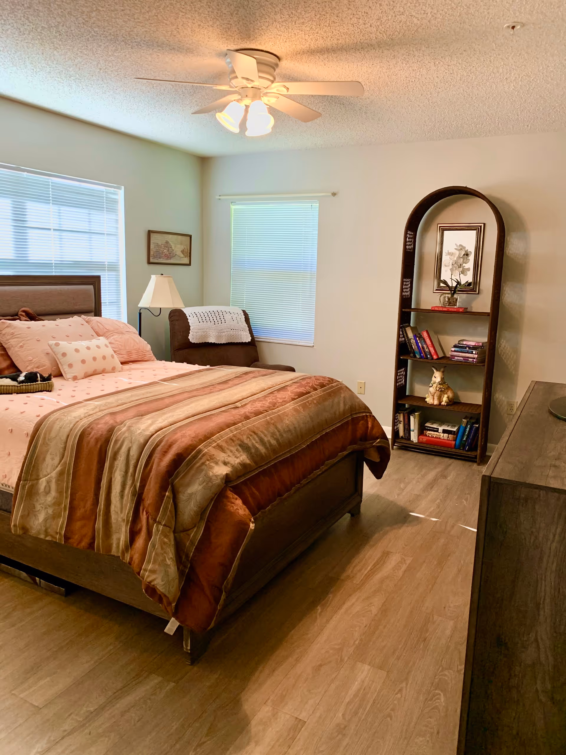 A tidy bedroom with a made bed, ceiling fan, armchair by two windows, and a curved bookshelf.