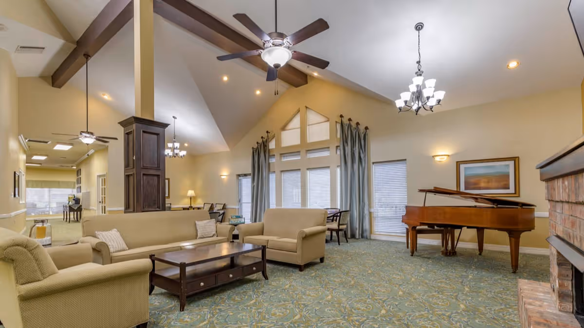 A spacious living room area with beige sofas and armchairs arranged around a dark wooden coffee table. The room features a high vaulted ceiling with exposed wooden beams, ceiling fans, and a chandelier. Large windows with curtains allow natural light to enter. A grand piano is positioned near a brick fireplace, and framed artwork hangs on the wall.