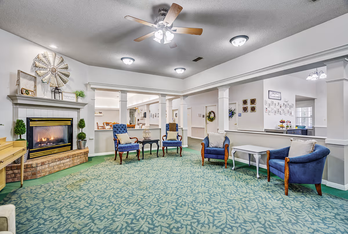 Spacious senior living common room with a lit fireplace, ceiling fan, patterned green carpet, and several blue upholstered chairs and tables.