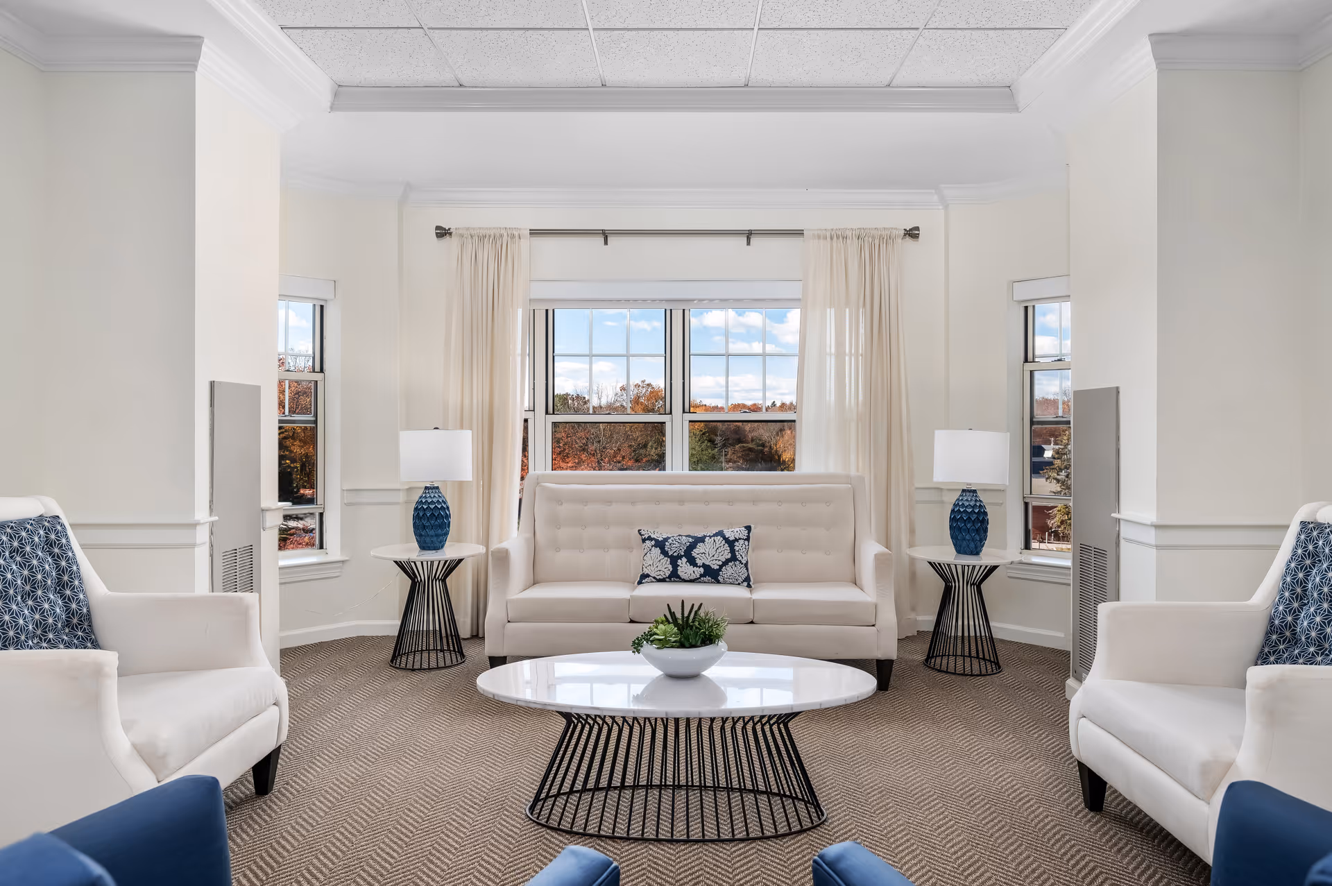 A bright and airy living room with a large window showing an outdoor view. The room features a white sofa with a blue and white decorative pillow, two white armchairs with blue patterned pillows, two side tables each with a blue lamp, and a round coffee table with a small plant. Light-colored curtains frame the window, and the walls are painted white.