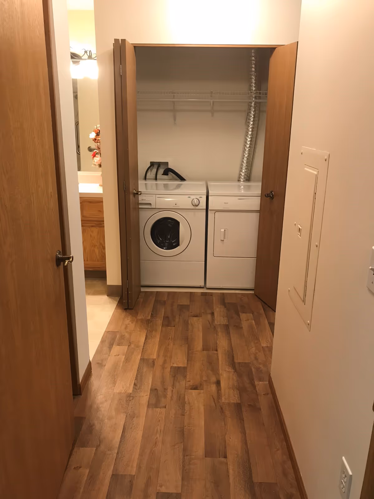 A hallway with wood flooring leading to a laundry closet with open wooden bi-fold doors revealing a white front-loading washing machine and a white dryer. To the left, there is a partial view of a bathroom with a sink, mirror, and floral arrangement.