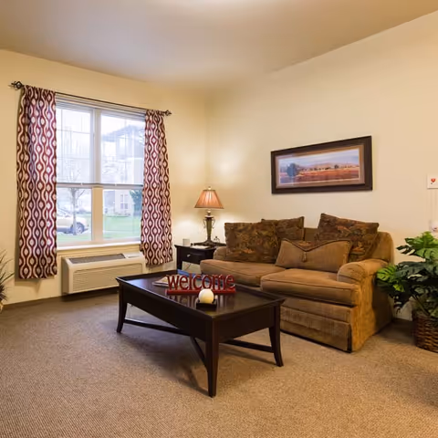 Cozy living room with a sofa, coffee table displaying a "welcome" sign, side table lamp, framed artwork, and a window with patterned curtains.