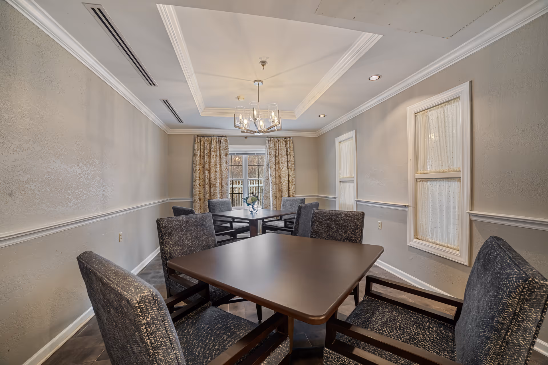 A dining room with two square dark wood tables, each surrounded by four upholstered chairs with armrests. The room has beige walls with white trim, two windows with sheer curtains on the right wall, and a larger window with patterned curtains at the far end. A modern chandelier hangs from the ceiling, which features crown molding and recessed lighting.