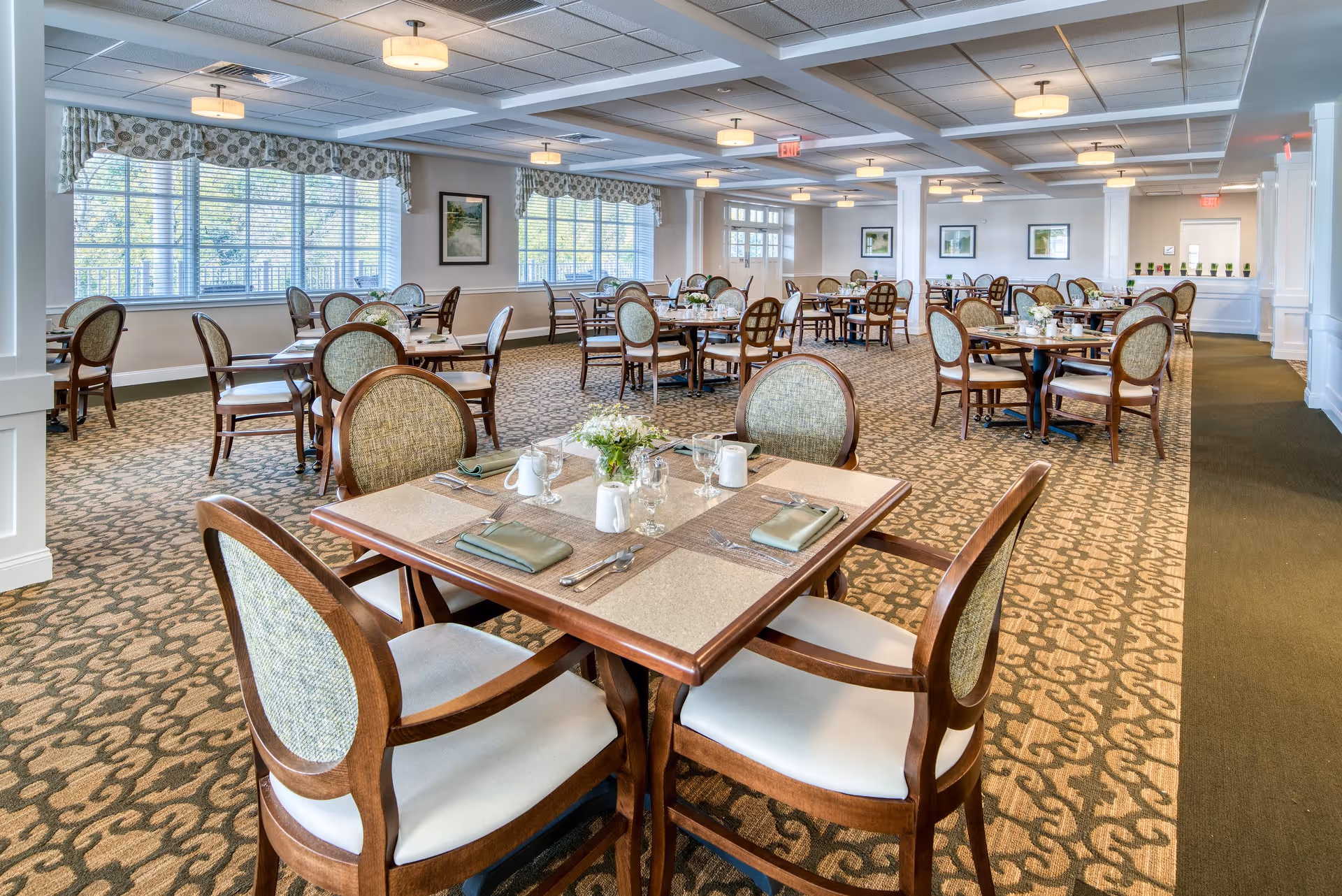 A spacious and well-lit dining room in a senior living facility with multiple wooden tables and cushioned chairs arranged neatly. Each table is set with placemats, napkins, glassware, and small floral centerpieces. Large windows with patterned valances allow natural light to fill the room, and framed artwork decorates the walls. The carpet has a decorative pattern, and ceiling lights provide additional illumination.