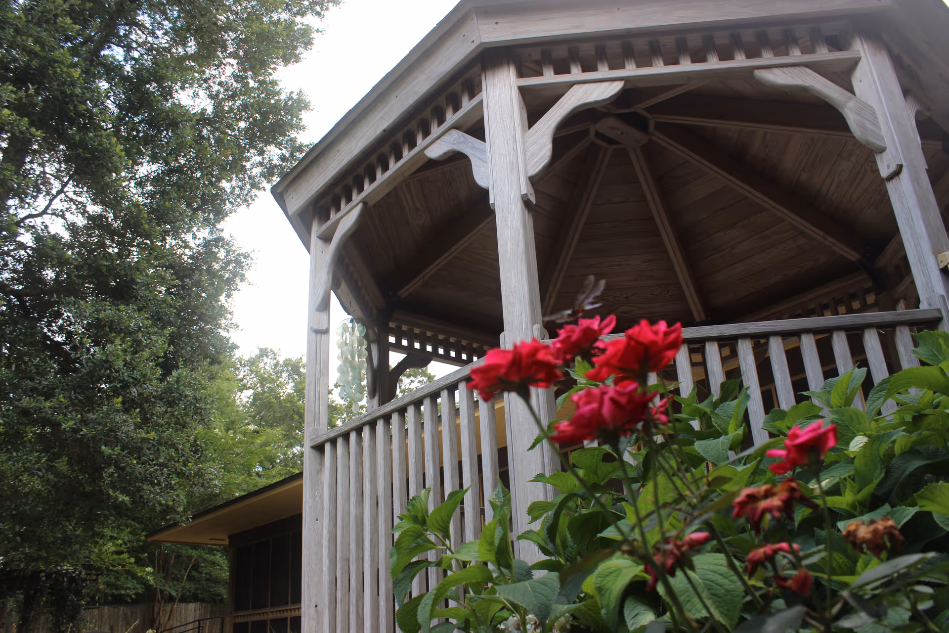 A wooden gazebo surrounded by green foliage and red flowers, with trees and part of a building visible in the background under a cloudy sky.