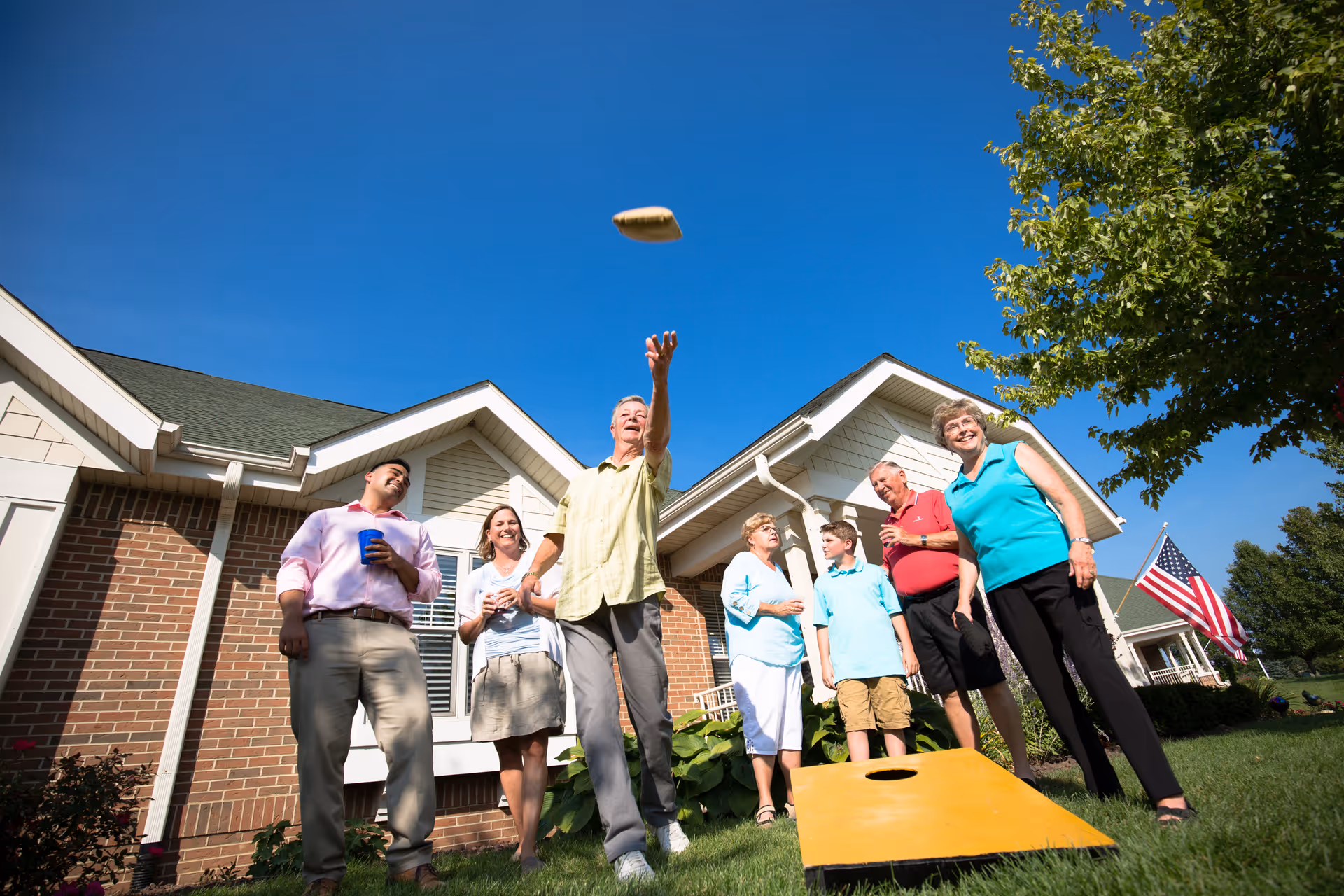 A group of six people, including seniors and a child, are playing cornhole outside on a sunny day in front of a brick and siding building with a green roof. One man is tossing a bean bag towards the cornhole board while others watch and smile. An American flag is visible in the background near a tree.