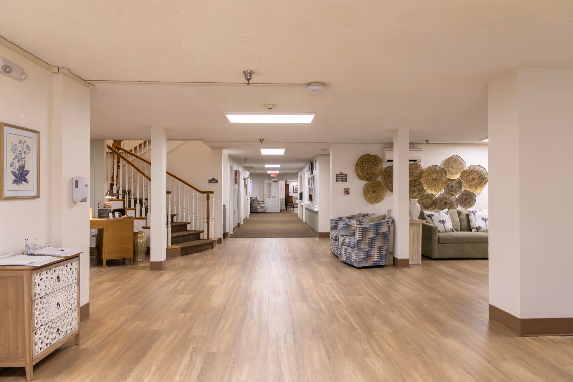 Interior view of a senior living facility hallway with wooden flooring, a staircase on the left, a seating area with patterned chairs and a couch on the right, and decorative wall art behind the seating area. The hallway extends into the distance with doors and lighting fixtures visible.