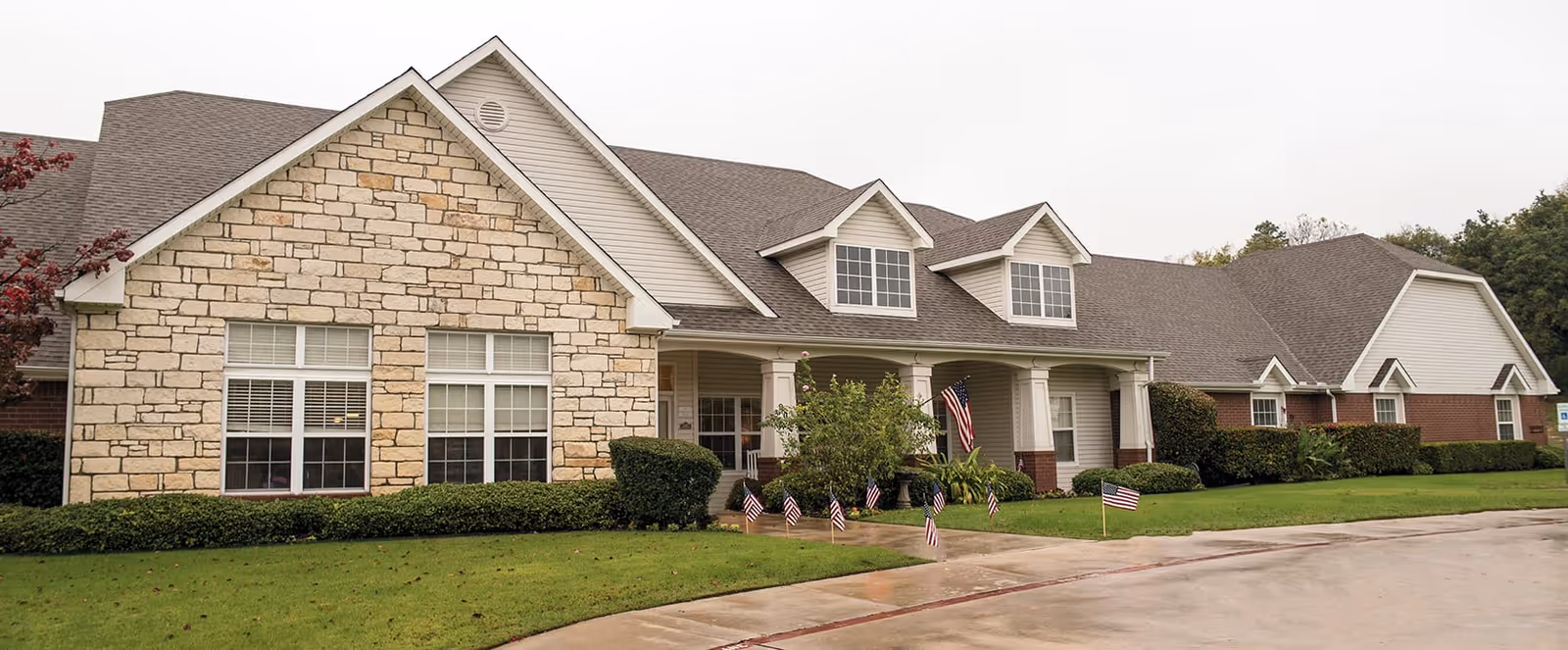 Exterior view of a single-story senior living facility building with stone and brick facade, multiple windows, a covered entrance with columns, and small American flags placed along the front lawn and walkway.