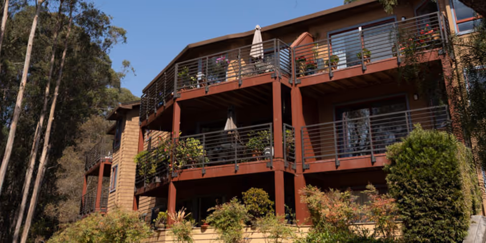 Exterior view of a multi-story residential building with balconies adorned with plants and outdoor furniture, surrounded by trees and greenery under a clear blue sky.