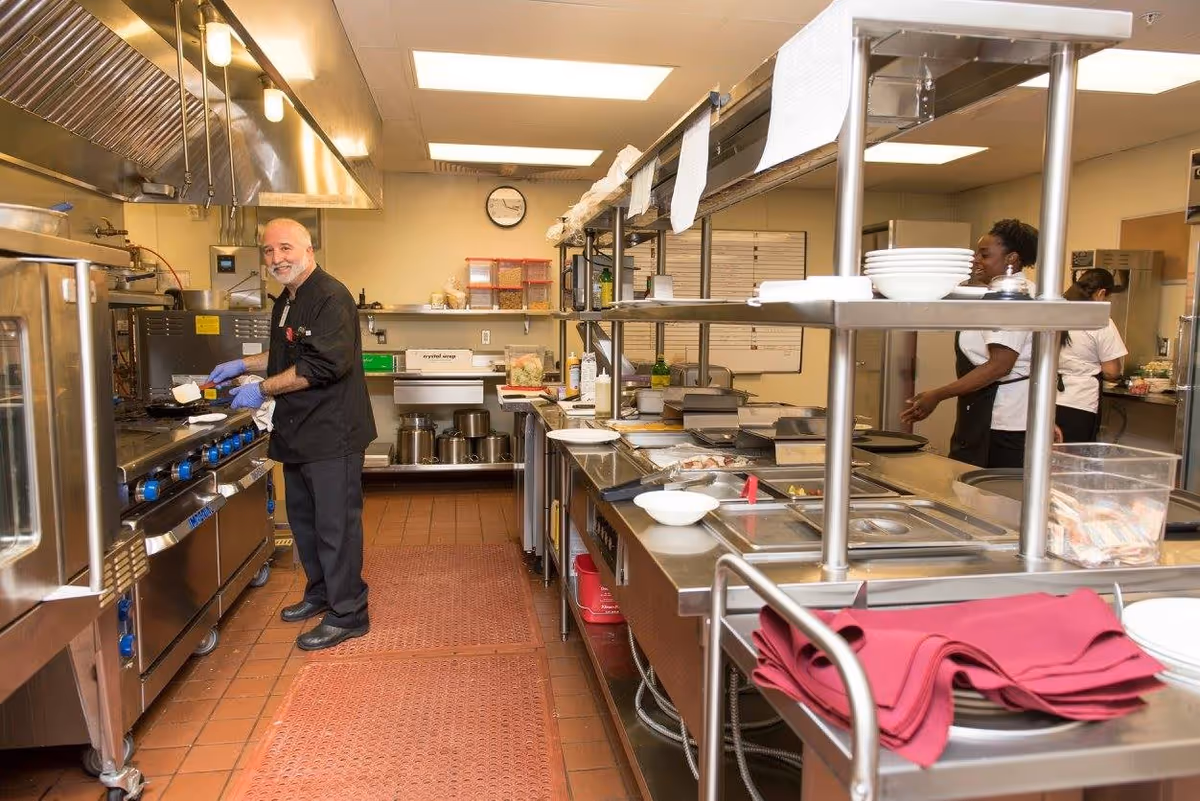 A commercial kitchen with a male chef cooking at the stove and two female kitchen staff working in the background. The kitchen has stainless steel counters, shelves with plates and bowls, and various kitchen equipment. The floor is tiled with red mats in front of the cooking area.