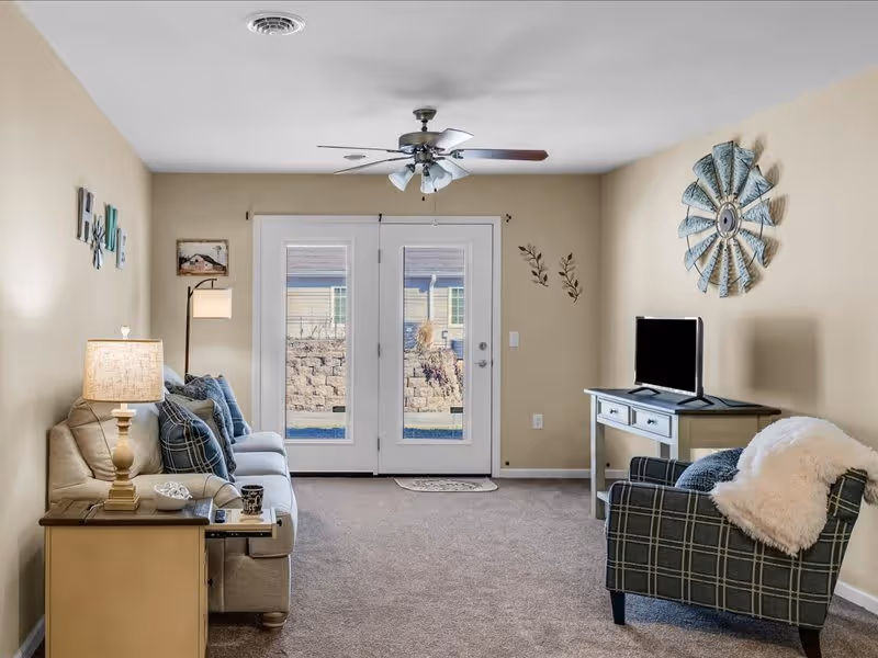 A cozy living room with beige walls and carpeted floor. There is a beige sofa with blue and plaid cushions on the left, next to a wooden side table with a lamp and decorative items. A plaid armchair with a white throw blanket is on the right, facing a small TV on a light-colored console table. French doors with glass panels lead outside, and a ceiling fan with lights is mounted on the ceiling. Wall decorations include a metal windmill and small framed pictures.
