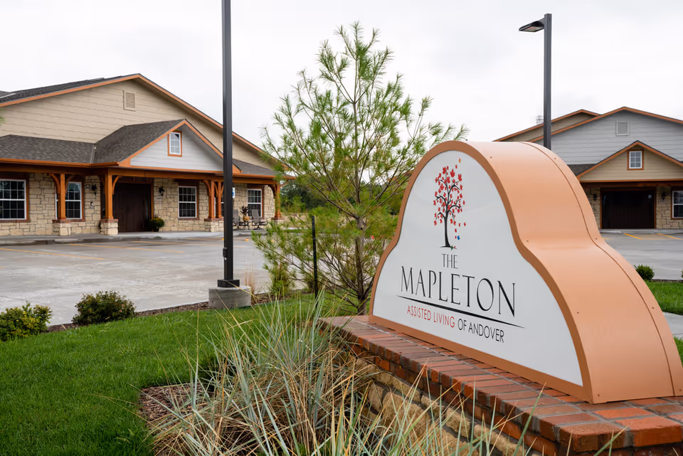 Exterior view of The Mapleton Assisted Living facility showing a large sign with the facility's name in front of a landscaped area with grass and plants. The building has a stone facade with wooden accents and a parking lot in front.
