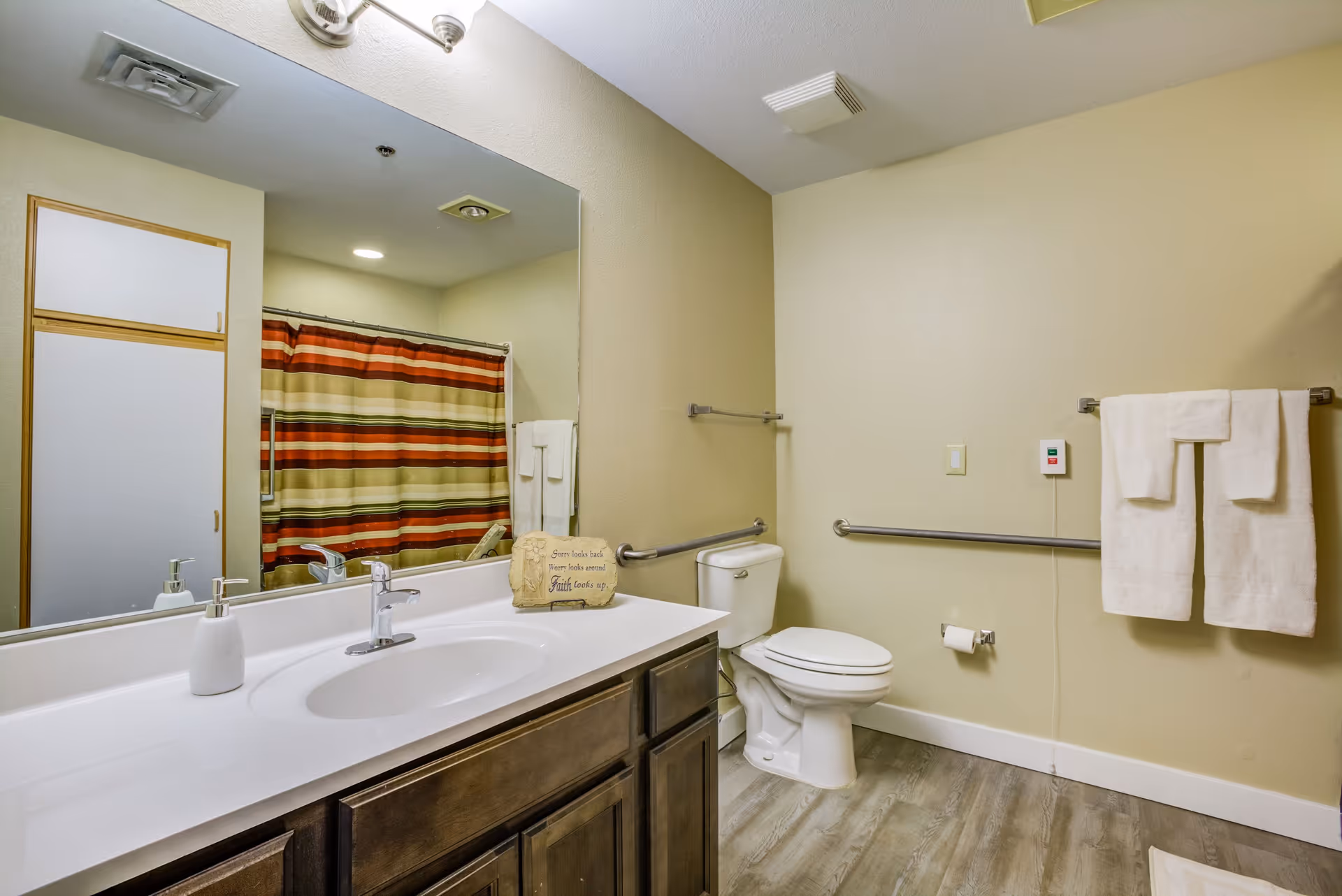 A clean and accessible bathroom with a white sink and countertop, dark wood cabinets, a large mirror, a toilet with grab bars on the walls, a striped shower curtain in shades of red, orange, and beige, and white towels hanging on a towel rack.