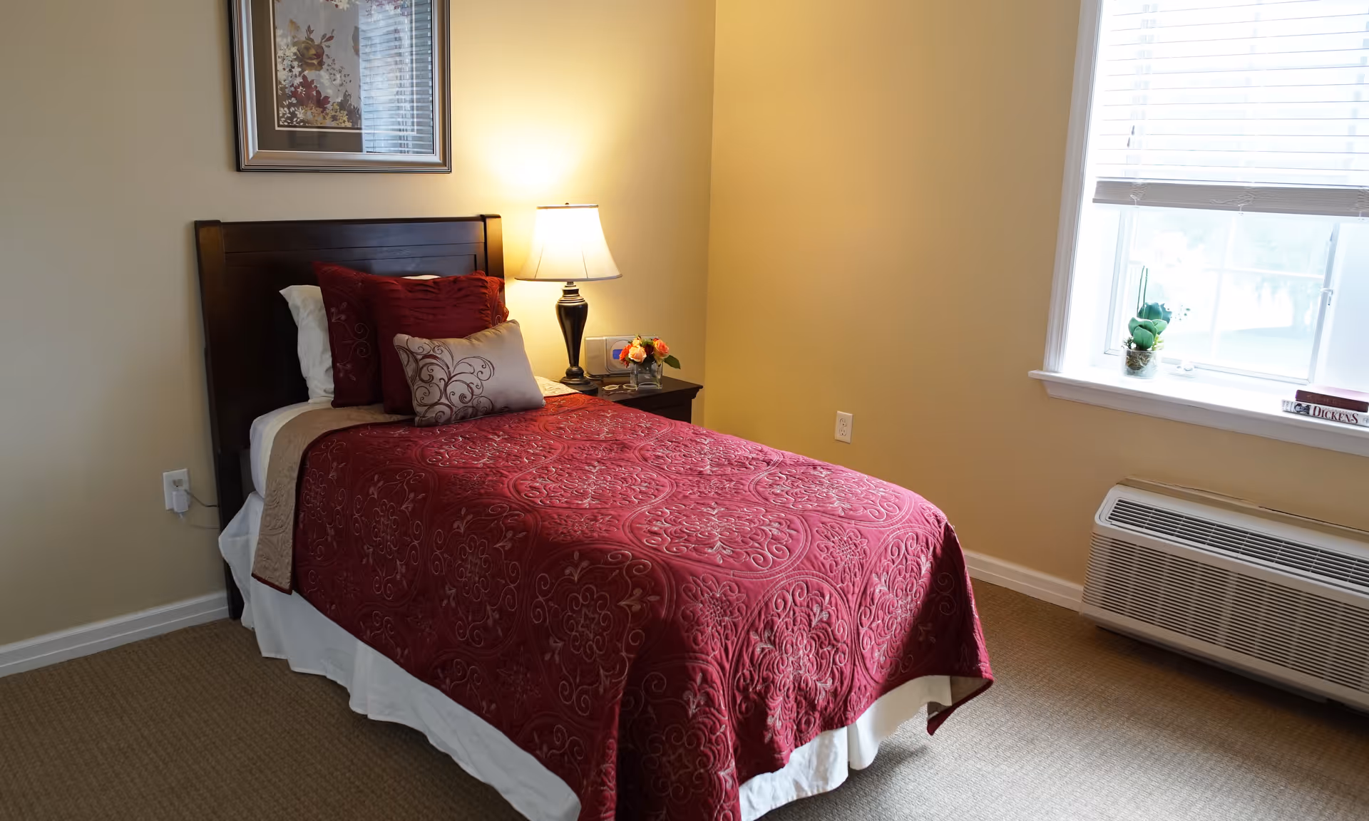 A neatly made single bed with a dark wooden headboard, covered with a red patterned bedspread and several decorative pillows. Next to the bed is a nightstand with a lamp, a small vase of flowers, and a clock. A window with white blinds lets in natural light, and a small plant and a book are placed on the windowsill. The room has beige walls and carpeted floor.