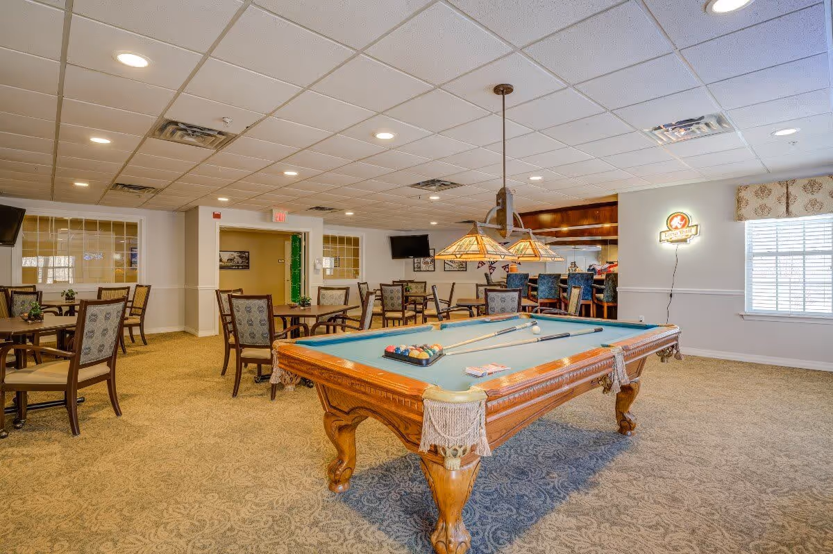 A spacious recreational room featuring a pool table with cues and balls set up, surrounded by several tables and chairs. The room has a carpeted floor, a ceiling with recessed lighting, and a bar area with high stools in the background. A window with blinds and a decorative valance lets in natural light, and a neon sign is mounted on the wall near the window.
