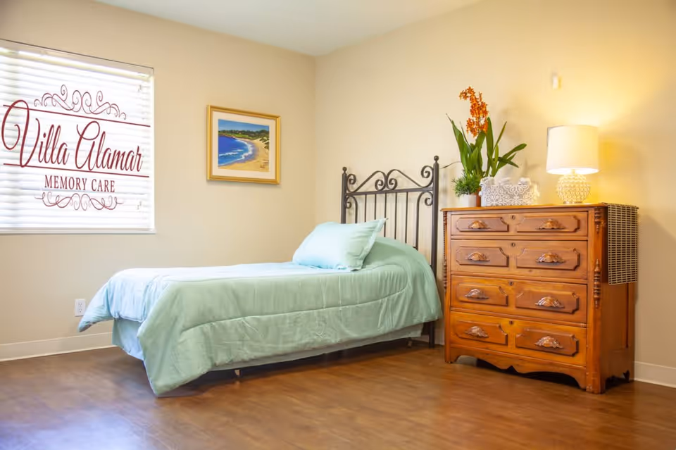A neatly made single bed with a light blue comforter and pillow in a room with wooden flooring. Next to the bed is a wooden dresser with multiple drawers, decorated with a potted plant, a tissue box, and a table lamp. A framed painting of a beach scene hangs on the beige wall above the bed. A window with blinds displays the text 'Villa Alamar Memory Care'.