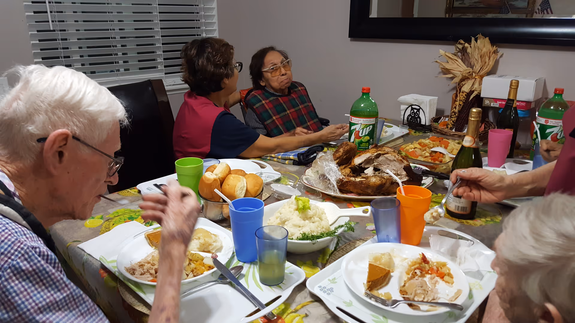 A group of elderly people sitting around a dining table enjoying a meal together. The table is set with plates of food including mashed potatoes, rolls, roasted turkey, vegetables, and slices of pie. There are colorful cups and bottles of 7UP on the table. The setting appears to be a cozy indoor dining room with a window and a large mirror on the wall.