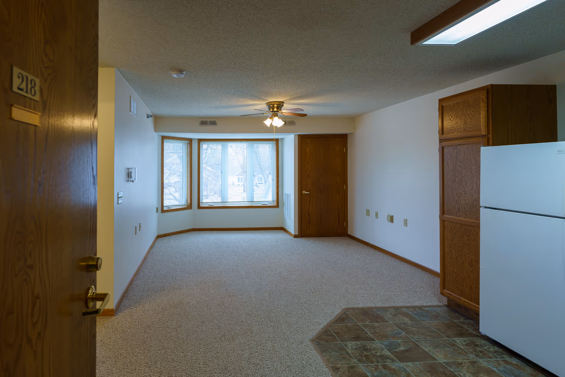 View of an empty apartment room with beige carpet and a ceiling fan with lights. There is a large window with a wooden frame at the far end, a wooden door to the right, and a white refrigerator next to wooden cabinets on the right side. The entrance door with the number 218 is partially visible on the left side.