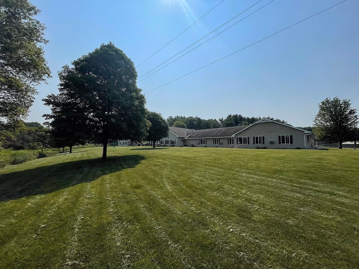 A large green lawn with neatly mowed grass and several trees casting shadows under a clear blue sky. In the background, there is a single-story building with multiple windows and a light-colored exterior, likely part of a senior living facility.