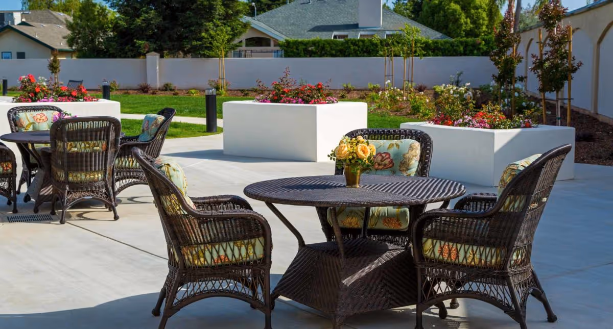 Outdoor patio area with wicker tables and chairs featuring floral cushions, surrounded by large white planters filled with colorful flowers and greenery, with a white wall and houses in the background under a clear sky.
